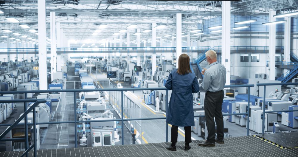Mature Engineer and Female Scientist Using Laptop Computer and Talking in a Factory Facility with Equipment Producing Modern Electronic Components for Different Industries