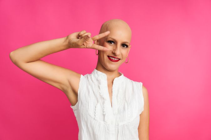 a woman suffering from cancer with a shaved head and a victory gesture on a pink background