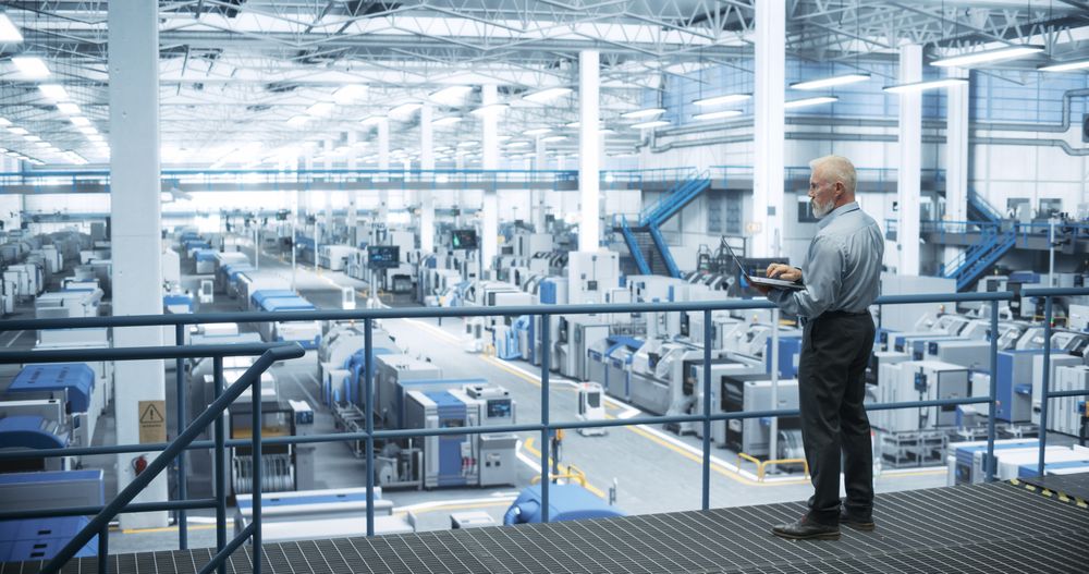 Experienced Male Engineer Standing on a Platform, Using Laptop Computer and Overlooking Production at a Modern Automated Electronics Manufacture with AI Support. Factory Executive at Work