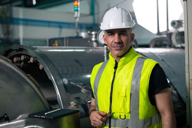 An industrial engineer wearing a safety helmet and reflective vest stands confidently in front of a machine in a factory. He is looking at the camera, representing professionalism and quality control