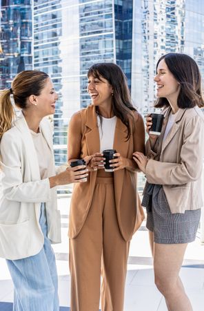 Three businesswomen are standing outside in front of a building, enjoying their office break, smiling and holding cups of coffee.