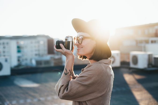 Chic woman with camera taking picture on rooftop at sunset