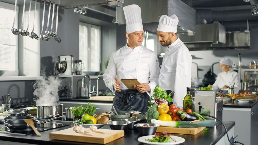 Two Famous Chefs Exchange Ideas About Video Blog Recipe they Watching on Tablet. They are Working in Big Restaurant Kitchen.