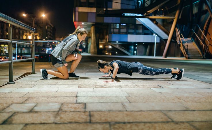 Woman doing push ups with support from personal trainer in the city at night