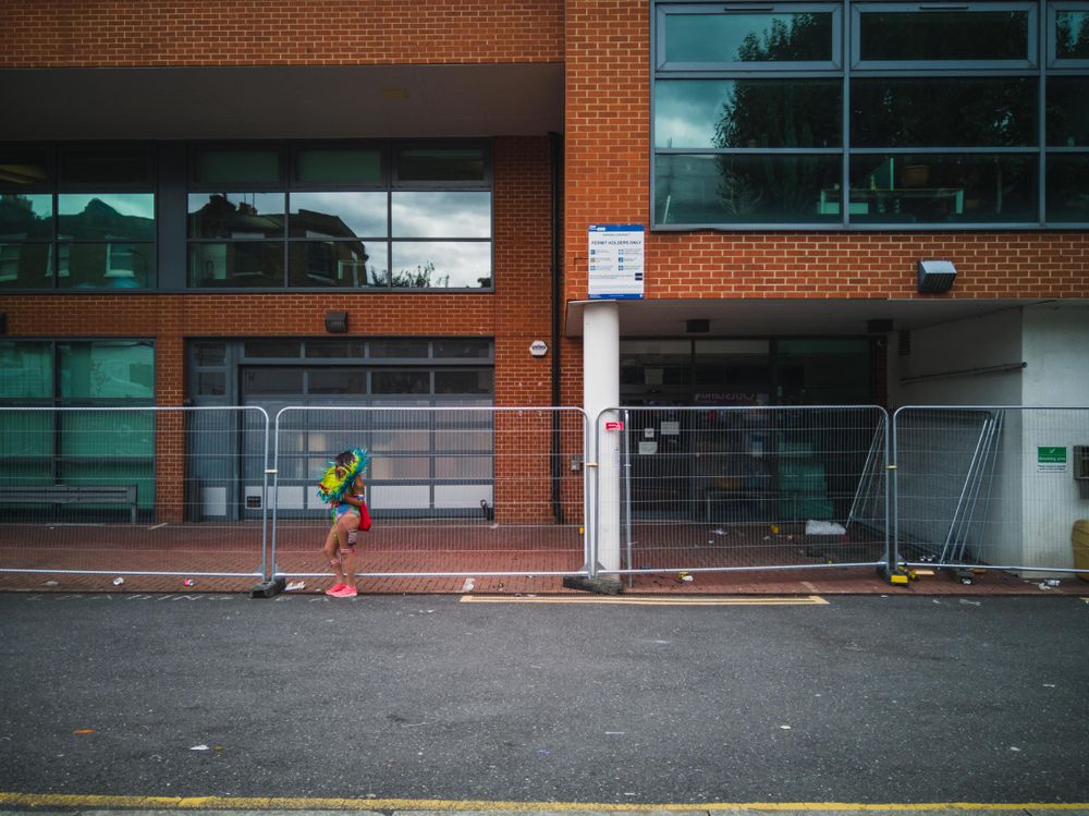 Young girl wearing colorful feathered ring around neck stands by fence on barren street after parade