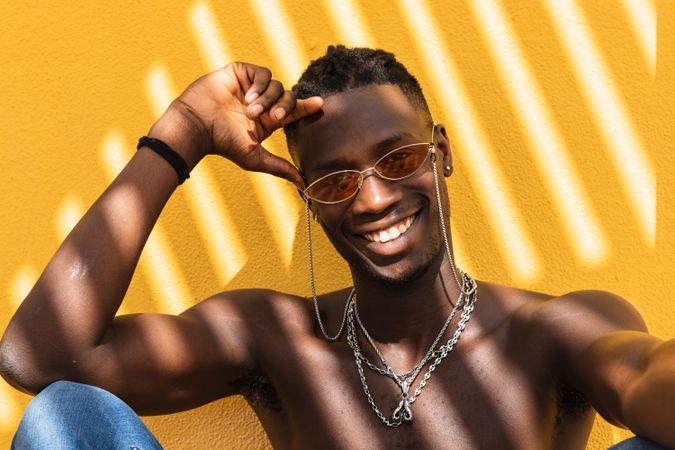 Cheerful Black man in trendy sunglasses sitting near wall