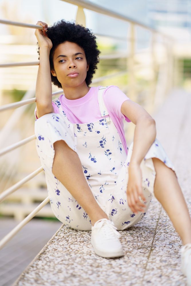 Woman in floral overalls sitting lost in thought leaning on handrail, vertical
