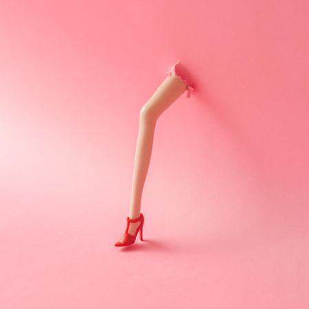 Doll leg on high heels breaking through pastel pink wall