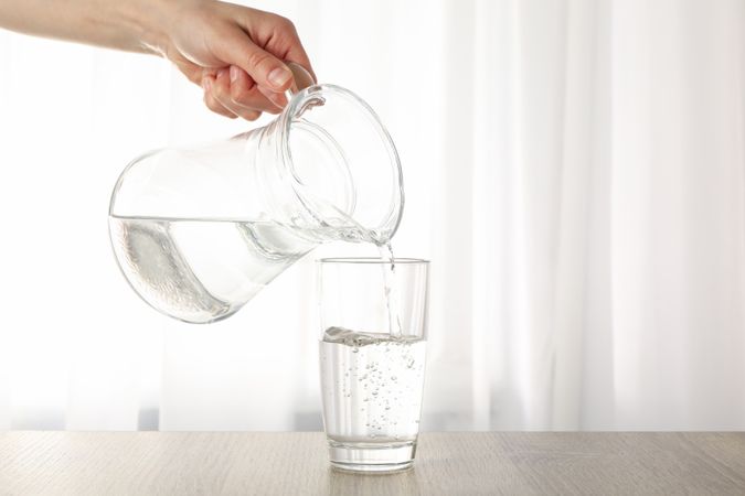 Hand pouring water into glass from pitcher in bright room near window