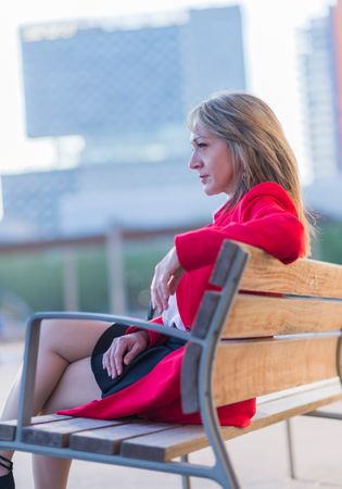 Side view of elegant woman in red jacket sitting on a bench outdoors while looking away in sunny day