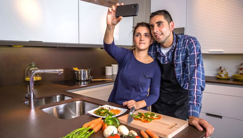 Content couple in a home kitchen taking a selfie with a smartphone while preparing meal