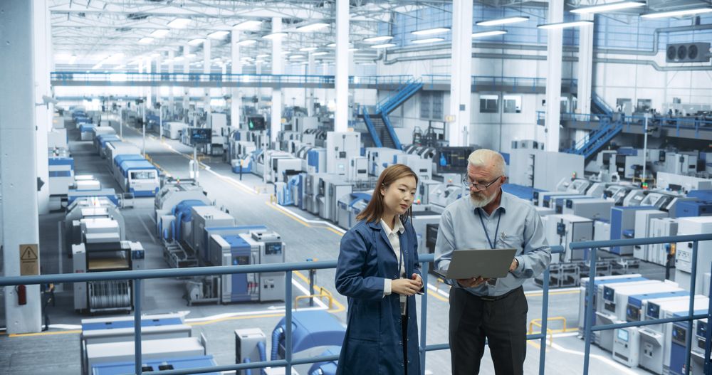 Portrait of a Mature Engineer and Positive Asian Scientist Using Laptop Computer and Talking in a Factory Facility with Equipment Producing Modern Electronic Components for Different Industries
