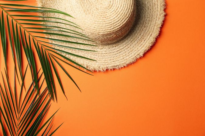 Straw hat and palm branches on orange background, top view