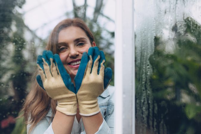 Smiling young woman inside greenhouse showing hand gloves