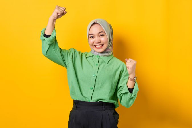 Smiling Muslim woman in headscarf and green blouse making two fists in victory