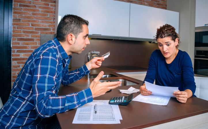 Man holding up credit cards discussing bills with partner in their kitchen