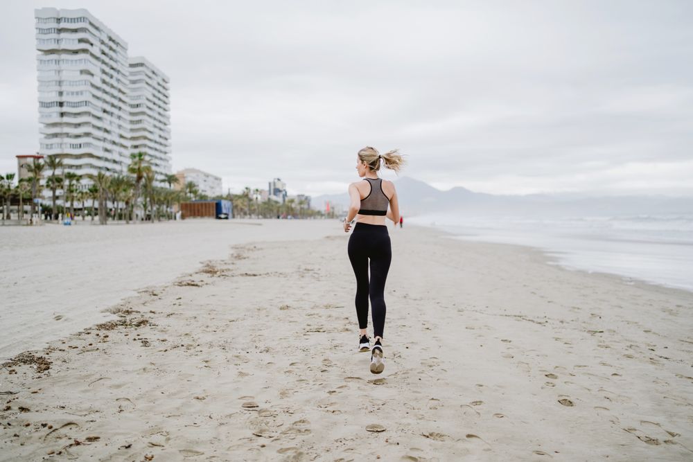 Female runner in stylish sportswear jogging along calm sandy seaside