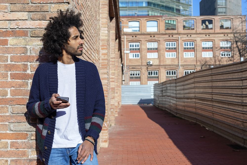 Black man using his smartphone with smile while leaning on a bricked wall outdoors on sunny day
