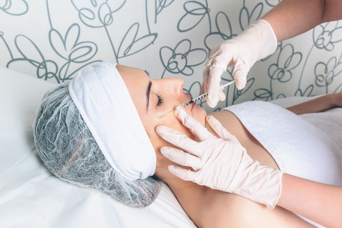 Side view of aesthetician's hand's in latex gloves injecting botox into female's cheek in a beauty salon