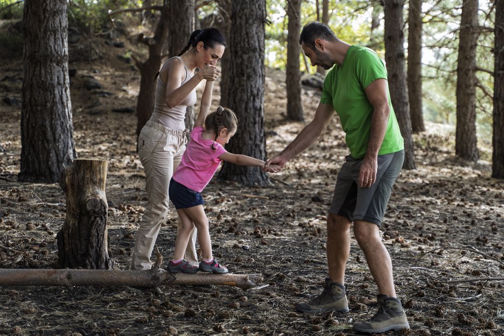 Parents helping daughter balance on log during forest walk adventure