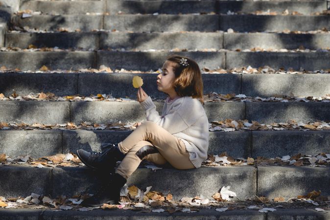 Child sitting on cement steps in outdoor park holding up a autumn leaf