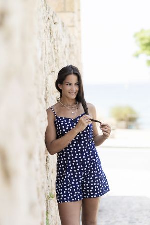 Latina woman in blue dotted dress leaning on wall outside and looking away playing with hair, vertical