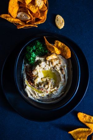 Looking down at creamy hummus dip in dark bowl with swirl of olive oil and chips
