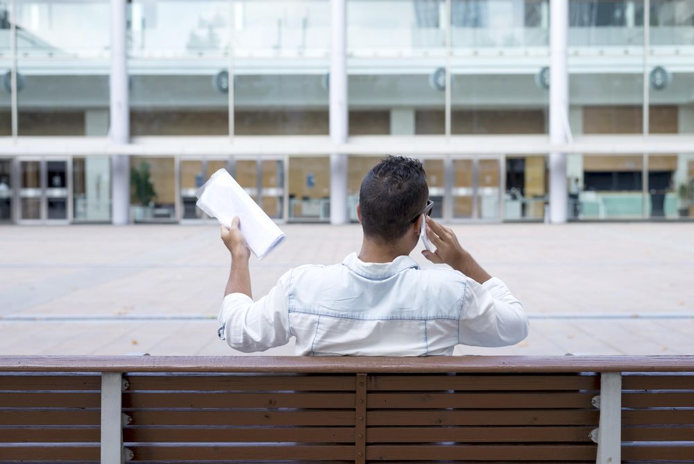 Back of male sitting on bench speaking on phone with documents in other hand