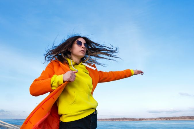 Trendy woman in sunglasses, orange coat, yellow hoodie and jeans in front of the bay