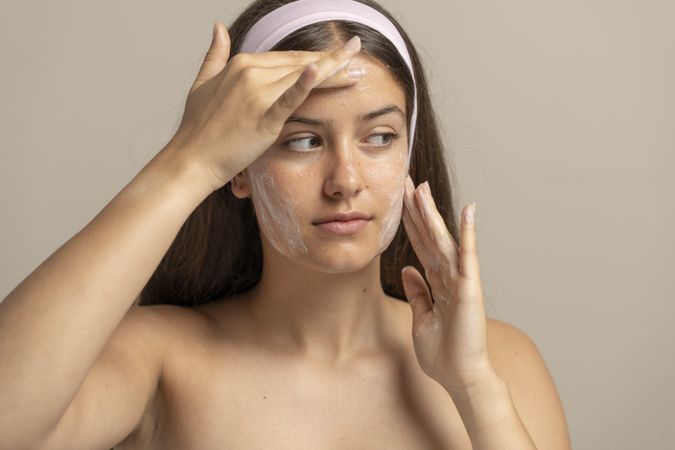 Young woman in studio gently applies white foam cleanser mask to her bare face, showcasing natural skin texture and visible pores in a realistic daily skincare ritual.