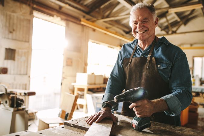 Happy mature male carpenter at his workshop holding power drill