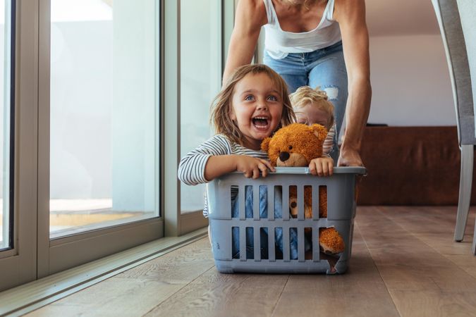 Excited little girl with teddy bear and her brother sitting in a laundry basket being pushed by mom