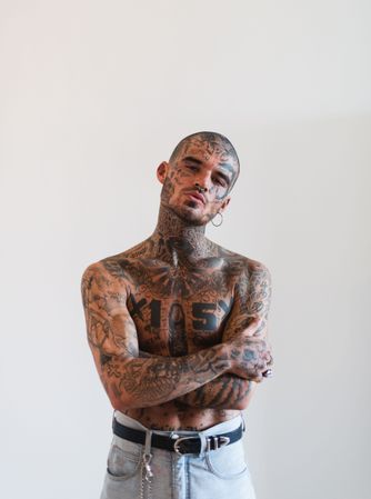 Shirtless Latino man in tough guy pose with full body and facial tattoos, shaved head and piercings