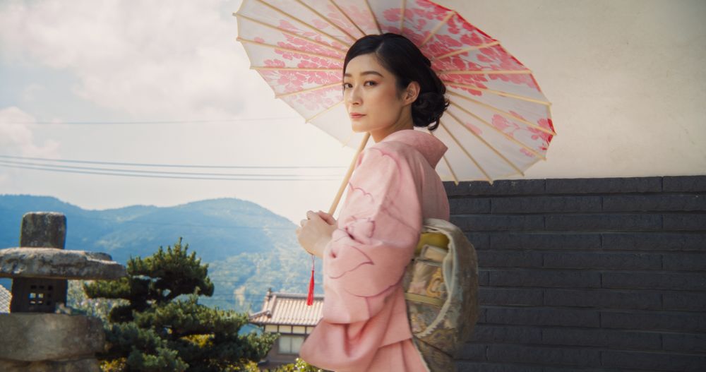 Young Japanese Woman in a Pink Kimono with Flowers and an Umbrella is Posing Outdoors.