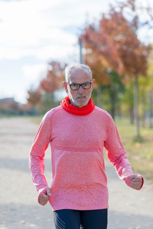 Grey haired man in red shirt and glasses jogging on in tree lined park