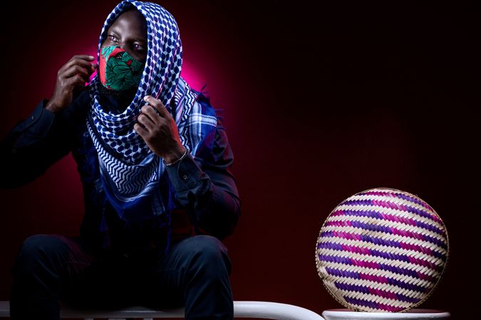 Man with facemask and headscarf sitting against red background