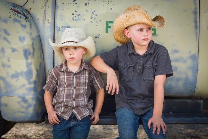 Two Young Boys Wearing Cowboy Hats Leaning Against an Antique Truck in a Rustic Country Setting