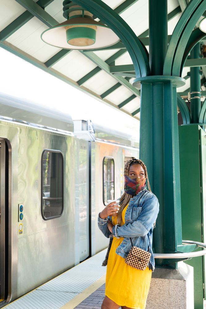 Woman standing waiting for city train wearing mask and holding coffee