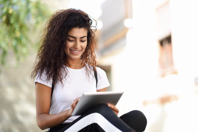 Happy Arab woman looking down at her digital tablet while sitting outdoors