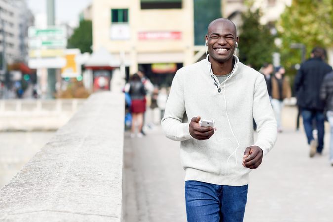 Smiling male with shaved head wearing casual sweater walking with headphones and phone