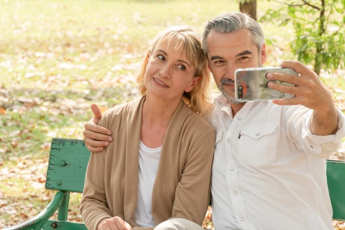 Male and female older couple taking selfie together on green park bench