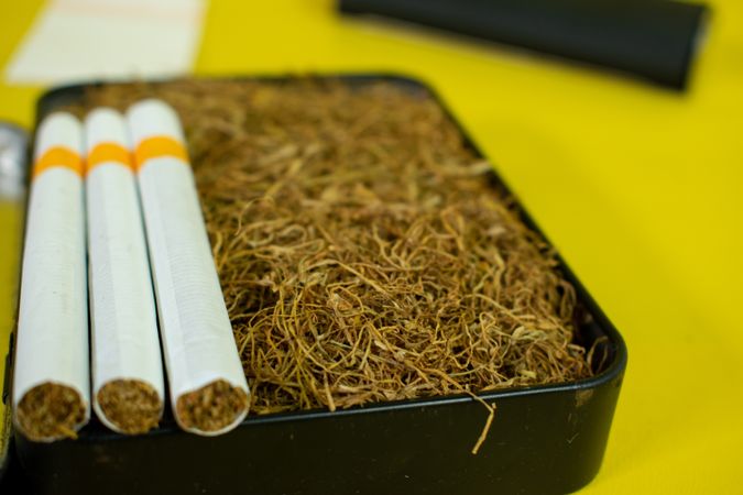 Close up of cigarettes resting on box of loose leaf tobacco on yellow table