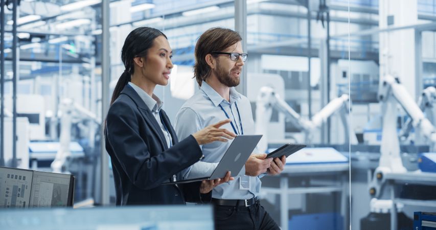 Factory Managers Using Computer Devices at a Modern Manufacture with Autonomous Robot Hands. Female and Male Specialists Monitoring Technical Data, Analyzing Reports and Planning Updates