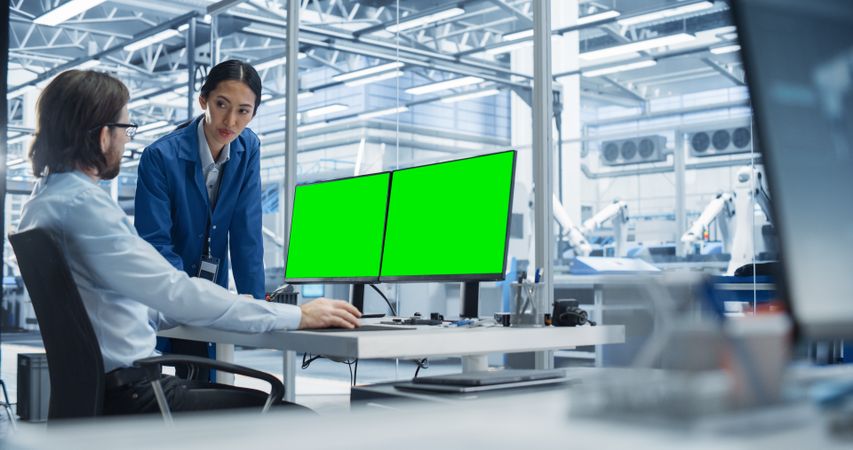 Portrait of a Young Male Engineer and Asian Team Leader Collaborating on a Project, Using Computer With Green Screen Chromakey On Display in a Factory with Equipment Producing Modern Technical Devices