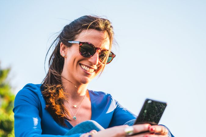 portrait of young woman with sunglasses using smartphone sitting on grass in park