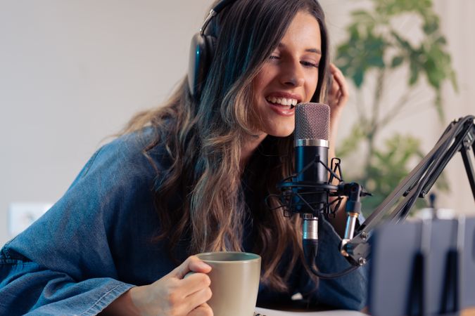 Young female podcast host enjoying coffee break during content recording session with professional microphone in creative home studio