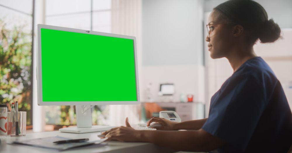 Portrait of a Black Female Medical Health Care Professional Working on Desktop Computer with Green Screen Mock Up Display in Hospital Office.