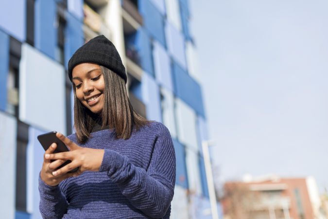 Female in hat and blue sweater checking cell phone outside blue building with space for text