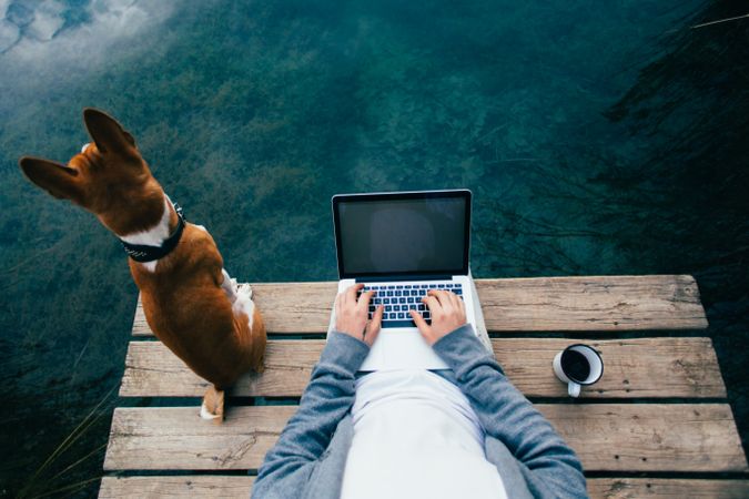 Man sitting on pier with coffee, dog and laptop