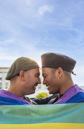 Gay couple under LGBT rainbow flag while laughing and looking at each other.